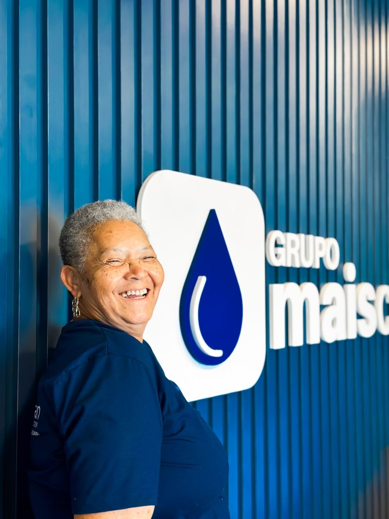 Colaboradora sorrindo ao lado da logo na sede do Grupo Mais Clean